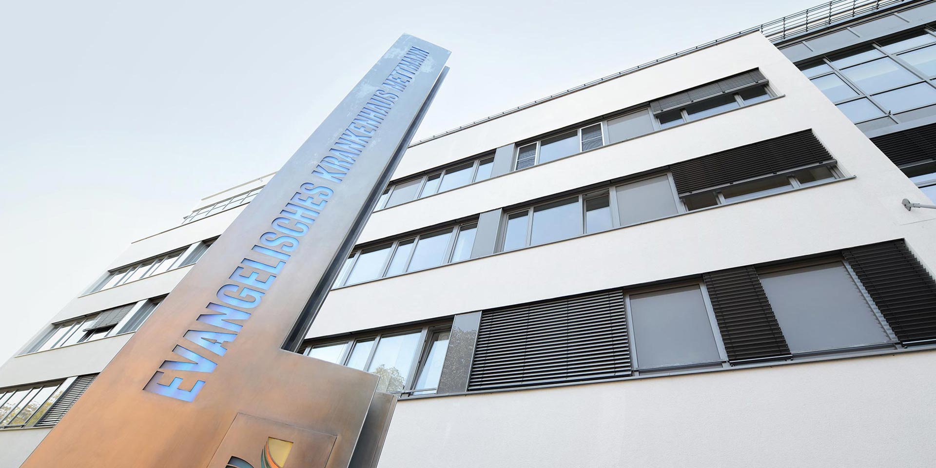 Außenansicht des Krankenhauses mit der Steele und dem Schriftzug Evangelisches Krankenhaus Mettmann – Blick Richtung Himmel.